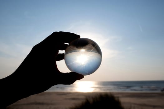 A hand of an  person holds a glass ball, the sea and the setting sun are reflected in the ball