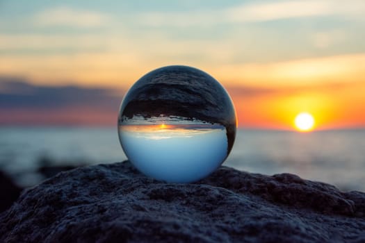 Glass ball on a rock at sunset on the beach, the sea and the setting sun are reflected in the ball