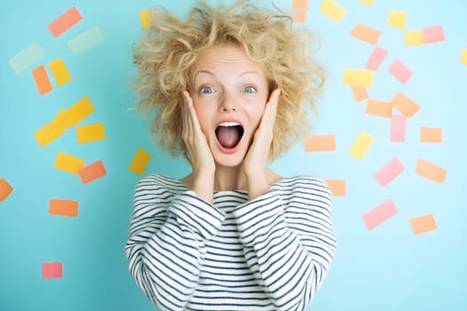 Woman with curly blonde hair expresses pure joy, holding her cheeks in excitement. She stands against a vibrant blue backdrop decorated with colorful paper scraps, creating a cheerful atmosphere.
