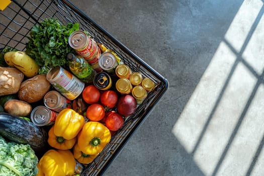 A shopping cart filled with fresh vegetables, fruits, and canned goods sits on the concrete floor of a grocery store, illuminated by natural light streaming through a window.