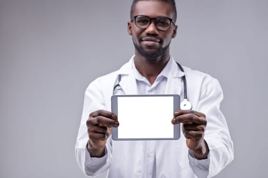 Smiling african american doctor wearing lab coat and stethoscope is holding a digital tablet with blank white screen, providing space for custom text or image
