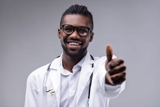 Young adult doctor, wearing a lab coat and stethoscope, smiling broadly while giving a thumbs up gesture, exuding confidence and positivity against a gray background