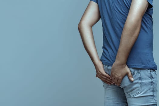 Man holding his bottom with both hands, experiencing discomfort from hemorrhoids, anal pain, and constipation, set against a soft gradient background