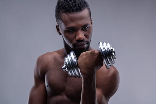 Muscular black man lifts a dumbbell with a determined expression, showcasing his strength and dedication to fitness against a gray studio backdrop