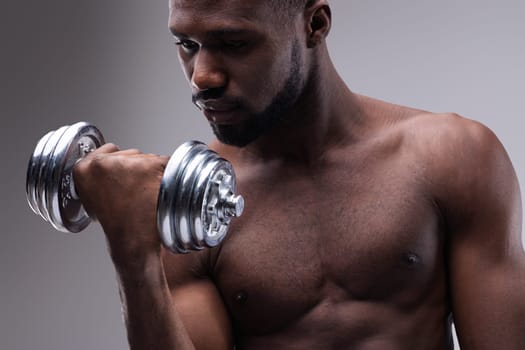 Strong young african american man lifting a dumbbell while intensely focusing on his arm, showcasing impressive muscles and strength during an energizing workout session