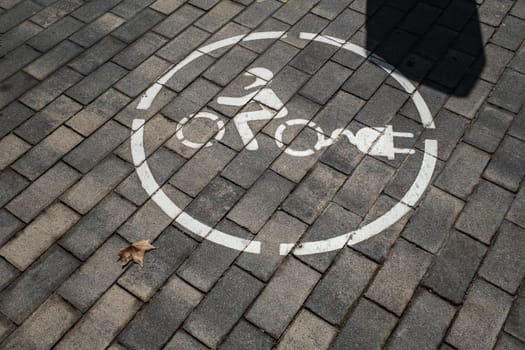 Service area for electric charging for motorcycles. Stylized drawing of a motorcyclist on two wheels with electric charging plug, circular outline, painted white. Background of concrete brick pavement.