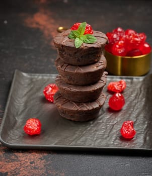 A stack of rich chocolate brownies topped with a glossy cherry and fresh mint leaf, standing on a dark background