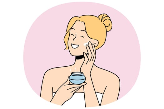Woman applies anti-aging cream to face to stay beautiful, standing in bath towel after shower. Happy blonde girl smiles and uses cosmetic cream to help prevent wrinkles and pimples.