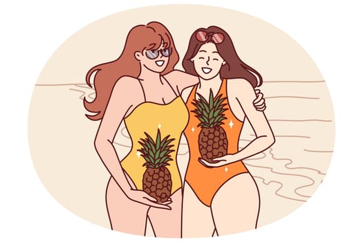 Two women in swimsuits stand on beach with pineapples in hands and smile inviting visit tropical island. Young happy girls in swimsuits spend holidays on sunny beach near sea or ocean