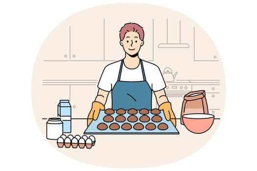 Man baker prepares oatmeal cookies or gingerbread while standing near kitchen table with ingredients. Baker guy dressed in apron and gloves proudly demonstrates dessert ready for baking.