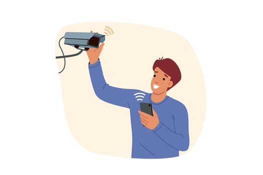 Man installs security camera on wall and uses mobile phone with wifi to monitor CCTV equipment. Happy guy with CCTV camera helping to secure house from illegal entry of thieves and criminals