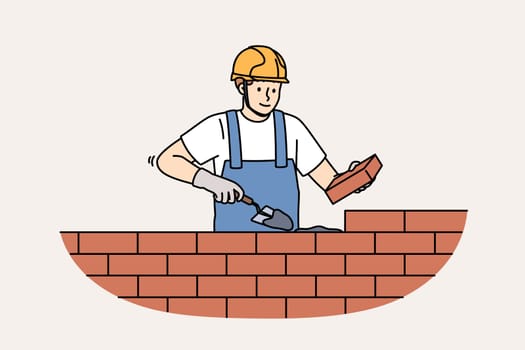 Man bricklayer builds brick wall by spreading concrete mortar to create strong house or cottage. Portrait of bricklayer in overalls and yellow helmet working in construction company