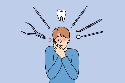 Man suffers from pain in teeth and appearance of caries, stands among dental instruments. Sick guy grabs face due to tooth pain and needs to visit dental clinic with professional doctors