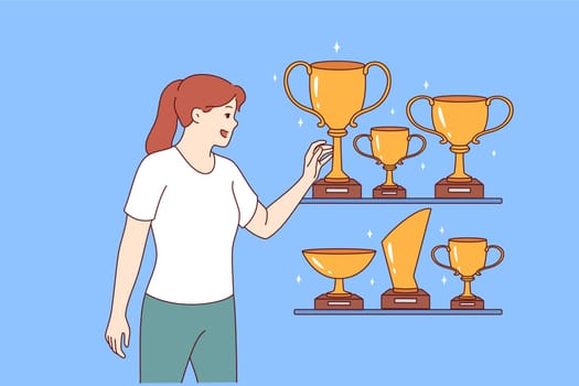 Woman leader stands near gold awards and cups won in sports competitions or professional competitions. Girl leader is proud to have trophies earned for outstanding results in business