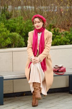 A woman sits comfortably on a park bench, dressed warmly in a beige coat, pink scarf, and tall brown boots while enjoying the autumn ambiance surrounded by greenery and fallen leaves.