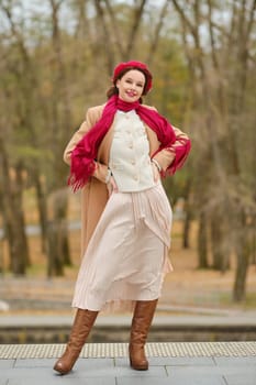 A fashionable woman stands playfully in a warm, stylish outfit featuring a scarf and long skirt. The autumn park setting adds to her charm as trees with colourful foliage surround her.