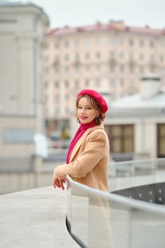 A woman stands on a balcony, dressed warmly in a beige coat with a pink beret and scarf. She gazes out thoughtfully over the cityscape, embracing the cool atmosphere of the day.