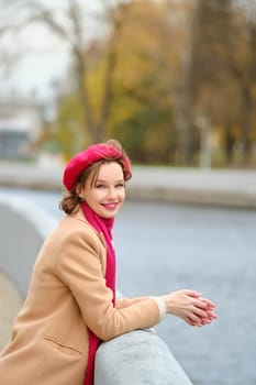 A joyful woman wears a red beret and beige coat while resting her hands on a concrete railing by the river.