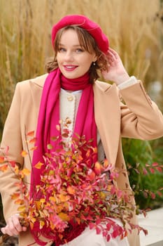 A woman dressed in a beige coat and pink scarf playfully poses outdoors. She holds a bouquet of vibrant autumn leaves, showcasing the beauty of the season against a natural backdrop.