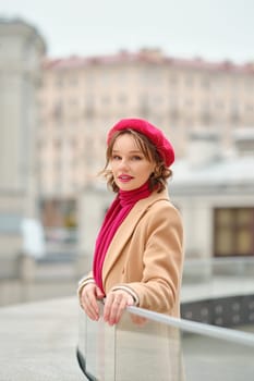 A stylish woman stands by a railing with a soft smile, wearing a red beret and matching scarf, against an urban backdrop.