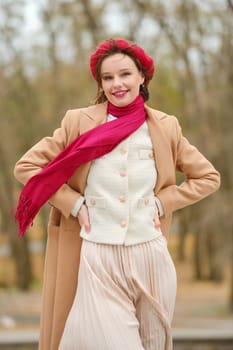 A smiling woman stands outdoors wearing a fashionable ensemble, including a light coat, scarf, and pleated skirt. She holds her hands on her hips against a backdrop of autumn trees.