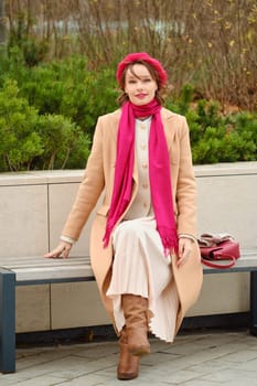 Cute woman sits comfortably on a bench in a park, dressed in a stylish beige coat, pink scarf, and hat, surrounded by greenery, evoking a cozy autumn atmosphere that highlights her fashion sense.