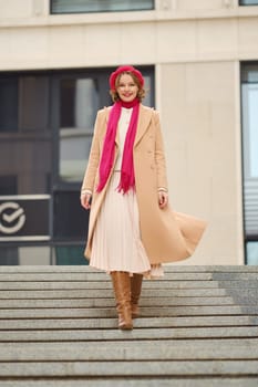 A young woman walks gracefully down stone steps wearing a chic long coat, scarf, and boots. The cold day enhances her radiant smile as she enjoys the vibrant atmosphere of the urban setting.