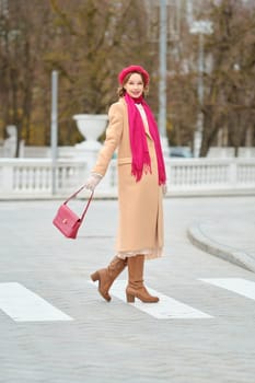 A fashionable woman is crossing the street in the city, wearing a beige coat, pink scarf, and red beret. She holds a bright pink handbag and walks confidently on the pedestrian crossing in autumn.