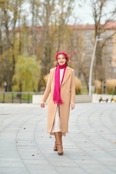 A woman walks through a picturesque park during autumn, dressed in a beige coat, scarf, and stylish boots. Her joyful expression complements the vibrant fall colors surrounding her.