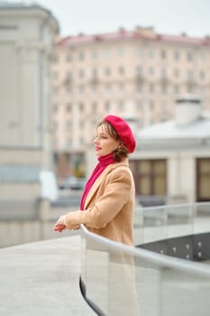 A woman wearing a red beret and matching scarf leans against a glass railing, gazing thoughtfully at the buildings in the background. The overcast sky adds a chilly atmosphere to the scene.