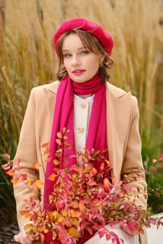A young woman stands in a park adorned in a warm coat, red beret, and scarf, holding vibrant autumn leaves. The golden backdrop of grass adds to the cozy fall atmosphere, reflecting seasonal beauty.