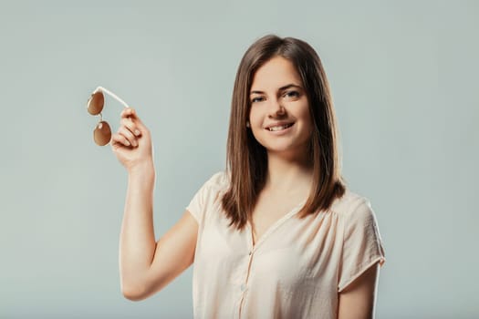 Studio shot featuring a beautiful young woman smiling brightly at the camera while holding a stylish pair of sunglasses against a soft gray background, exuding confidence and joy