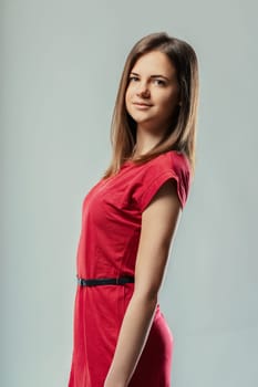 Studio portrait of a beautiful young woman with long brown hair wearing a stylish red dress and posing confidently against a neutral gray background