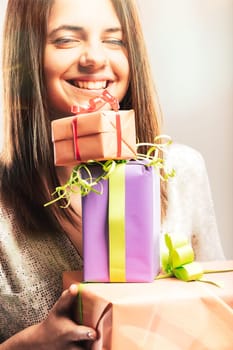 Young adult woman holding a stack of colorful wrapped gifts with ribbons and bows, smiling at the camera with a look of happiness and gratitude