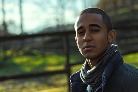 Young adult man wearing a scarf and jacket, enjoying the sunlight in a natural environment, possibly a park or countryside, with a blurred background suggesting trees and a fence