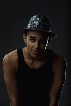 Young man wearing a stylish hat, striking a pose for a dance photoshoot in a studio featuring a striking black background. Portrait
