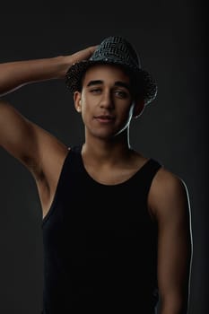 Portrait of a young fashionable man with hat posing on dark background, looking at camera. Portrait