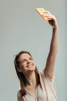 Cheerful young woman holding her smartphone high, capturing a selfie while looking up and smiling joyfully, radiating positivity and happiness against a gray studio backdrop