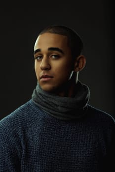 Portrait of a serious young man wearing a winter sweater and scarf, looking intensely at the camera against a dark background. Portrait