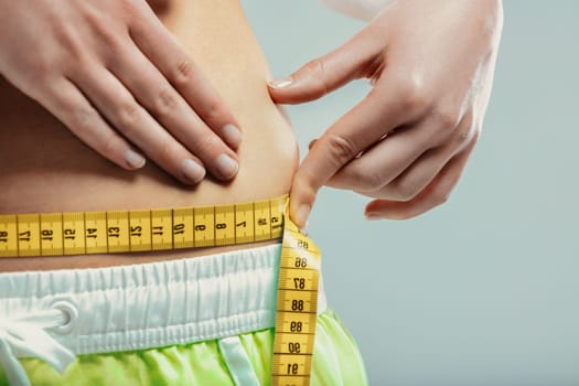 Close-up of a woman pinching her belly fat while measuring her waist with a yellow tape measure, expressing concerns about weight gain and body image