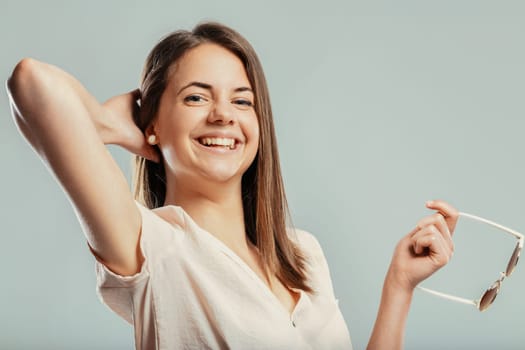 Cheerful young woman holding sunglasses and smiling with her hand behind her head, radiating happiness and relaxation, capturing a moment of carefree joy and positivity