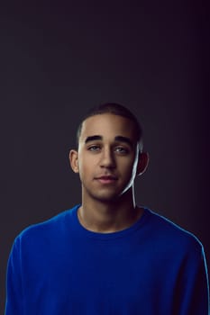 Portrait of a young man wearing a blue sweater, posing confidently while gazing directly at the camera against a dark background. Portrait