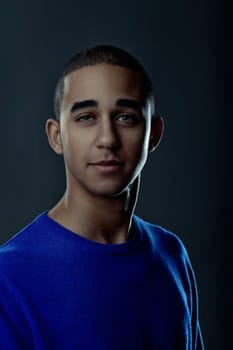 Portrait of a confident young man with short hair, wearing a blue sweater, posing against a dark studio backdrop. Portrait