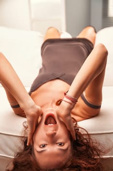 Young woman with curly red hair lying upside down on a white sofa, hands on her face, joyfully shouting and expressing excitement while looking directly at the camera