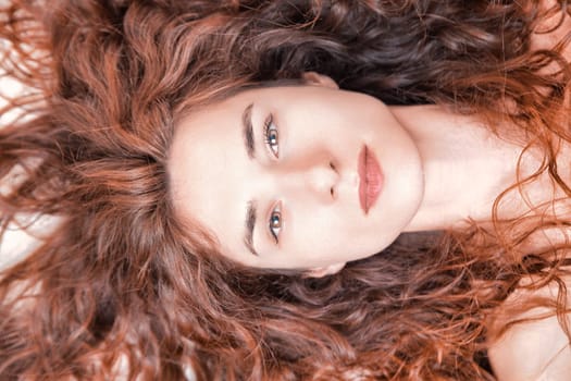 Close-up studio portrait featuring a stunning young woman with long, curly red hair, lying down and gazing at the camera with a serene and peaceful expression