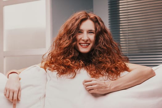 Young cheerful woman with long, curly red hair is smiling and relaxing on a comfortable white sofa in a cozy living room, enjoying a moment of peace and happiness