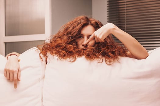 Playful young woman with vibrant red curly hair is lying on a comfortable sofa, her face partially hidden by the cushions as she points her finger at her nose and makes a silly expression