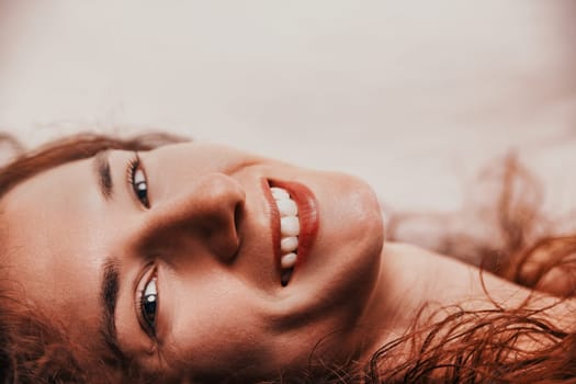 Close-up portrait featuring a cheerful young woman with long red hair lying down and smiling brightly, showcasing her teeth and exuding a sense of joy and contentment