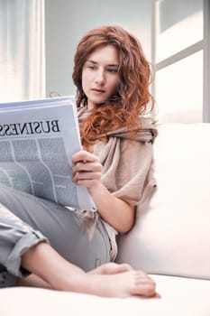 Young redhead business woman sitting barefoot on a white sofa at home, engrossed in reading the latest financial news from a business newspaper, enjoying a moment of leisure and relaxation