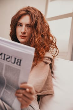Young woman with curly red hair sitting comfortably in bed, engrossed in reading the business section of a newspaper, catching up on current events and financial markets